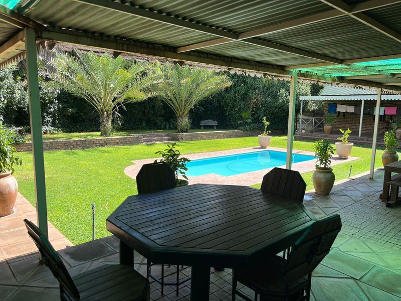 A covered outdoor patio area overlooking a swimming pool and landscaped garden with mature palm trees and brick retaining walls.