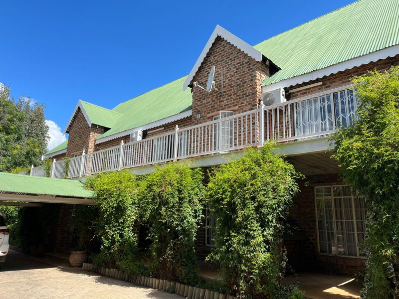 A two-story face-brick building featuring a green corrugated roof, an upper-level balcony with white balustrades, and a paved driveway with a covered carport.