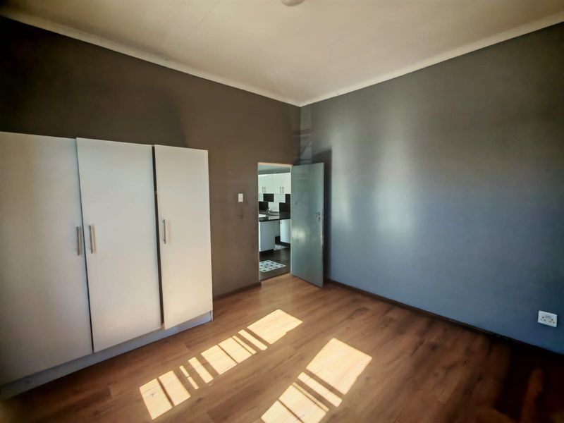 A spacious bedroom featuring laminate flooring, a built-in wardrobe, and neutral grey walls, with a view into the adjacent kitchen area.
