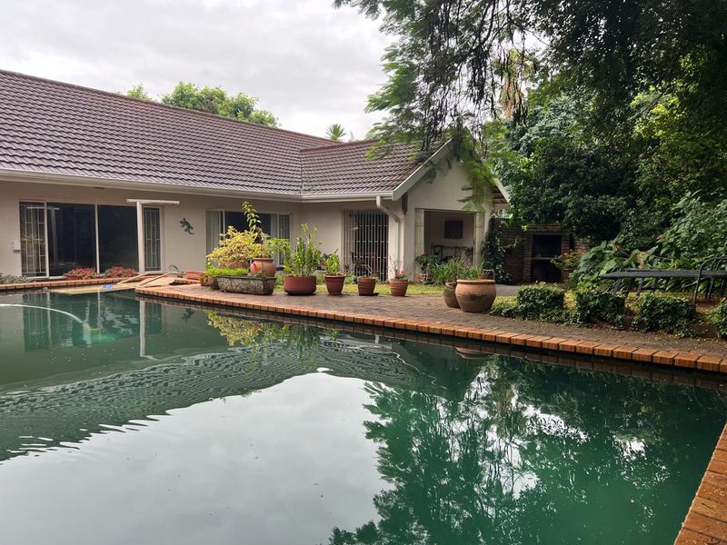 Rear view of the property featuring a swimming pool, brick patio area, and a built-in outdoor braai, surrounded by mature garden landscaping.