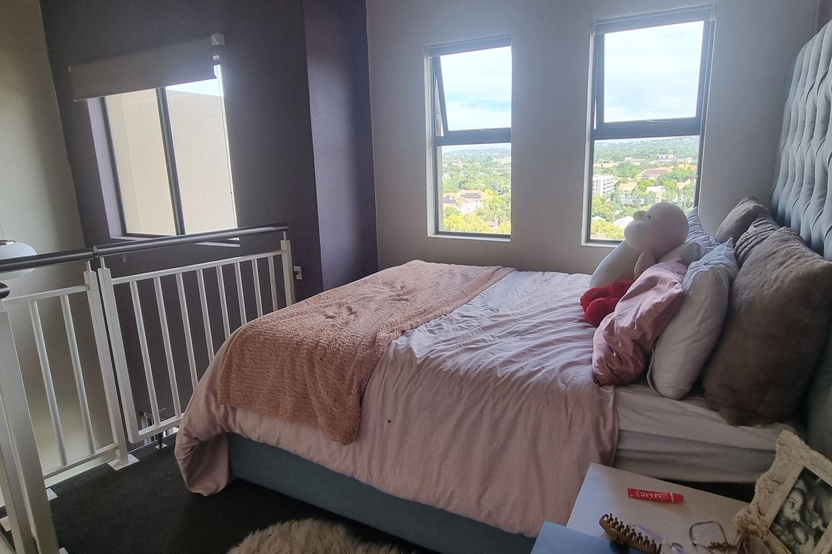 A loft-style bedroom featuring an exposed brick wall, metal balustrade overlooking the lower level, and large windows providing natural light and elevated views.