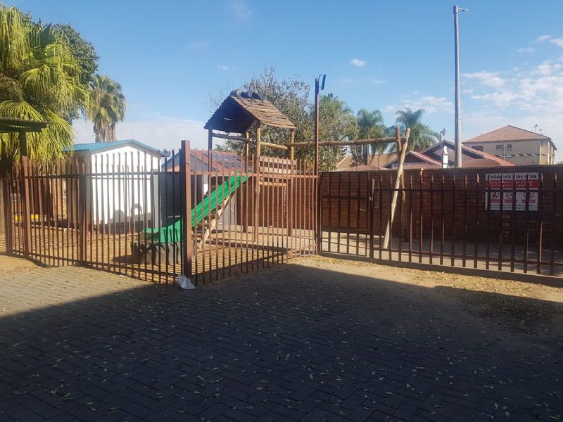 A communal outdoor area featuring a paved surface, metal security fencing, and a children's play structure within a residential complex.