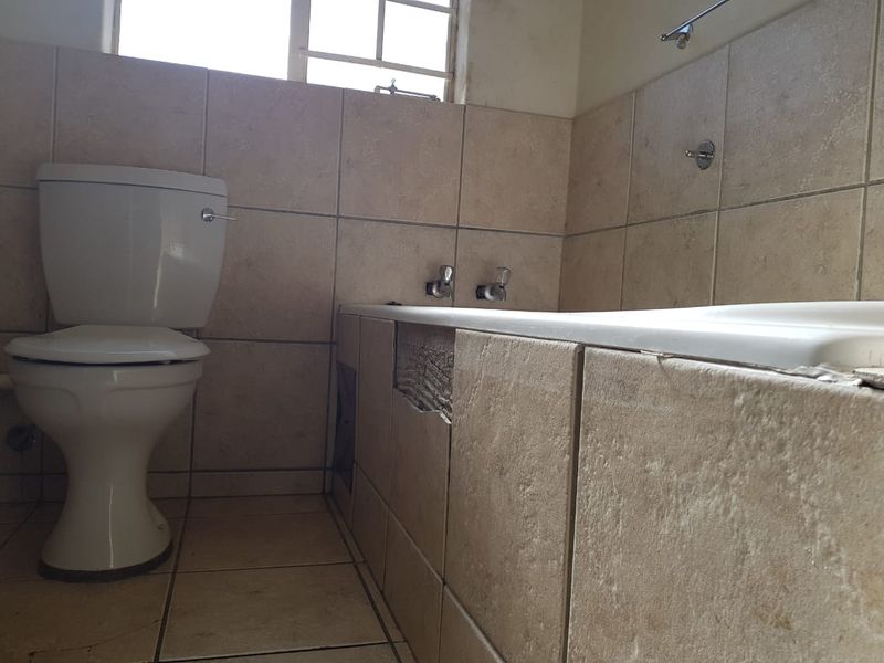 A bathroom featuring a toilet and bathtub with tiled finishes, currently requiring repairs to the bathtub apron and general maintenance.