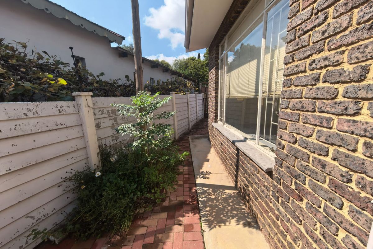A side exterior view of the townhouse featuring a brick facade, paved walkway, and a boundary fence with visible signs of weathering.