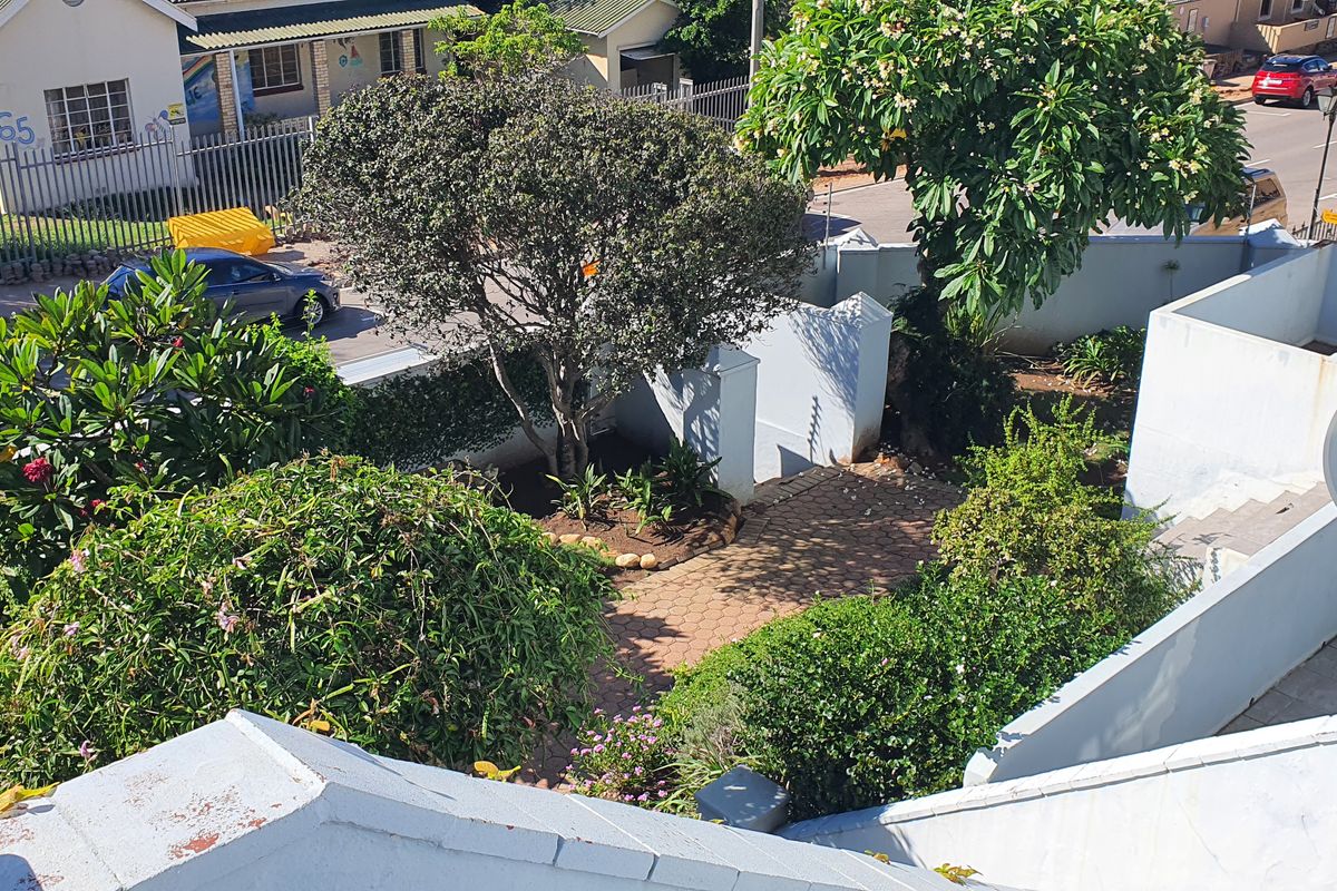 An elevated aerial view of the property grounds, featuring a paved walkway, established garden landscaping, and boundary walling.