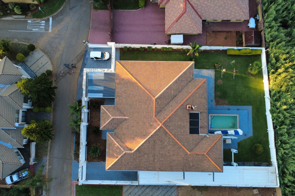 Aerial view of the property showcasing the tiled roof, swimming pool with solar heating, surrounding garden, and paved driveway area.