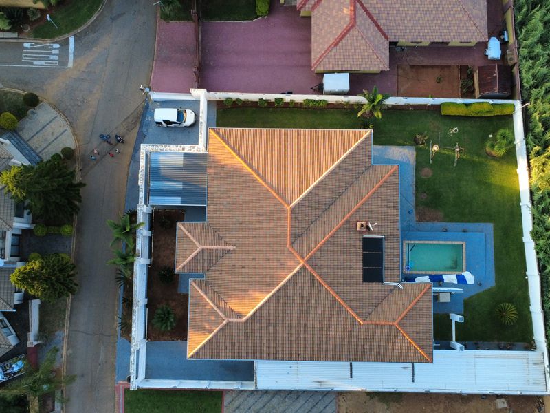 Aerial view of the property showcasing the tiled roof, swimming pool with solar heating, surrounding garden, and paved driveway area.