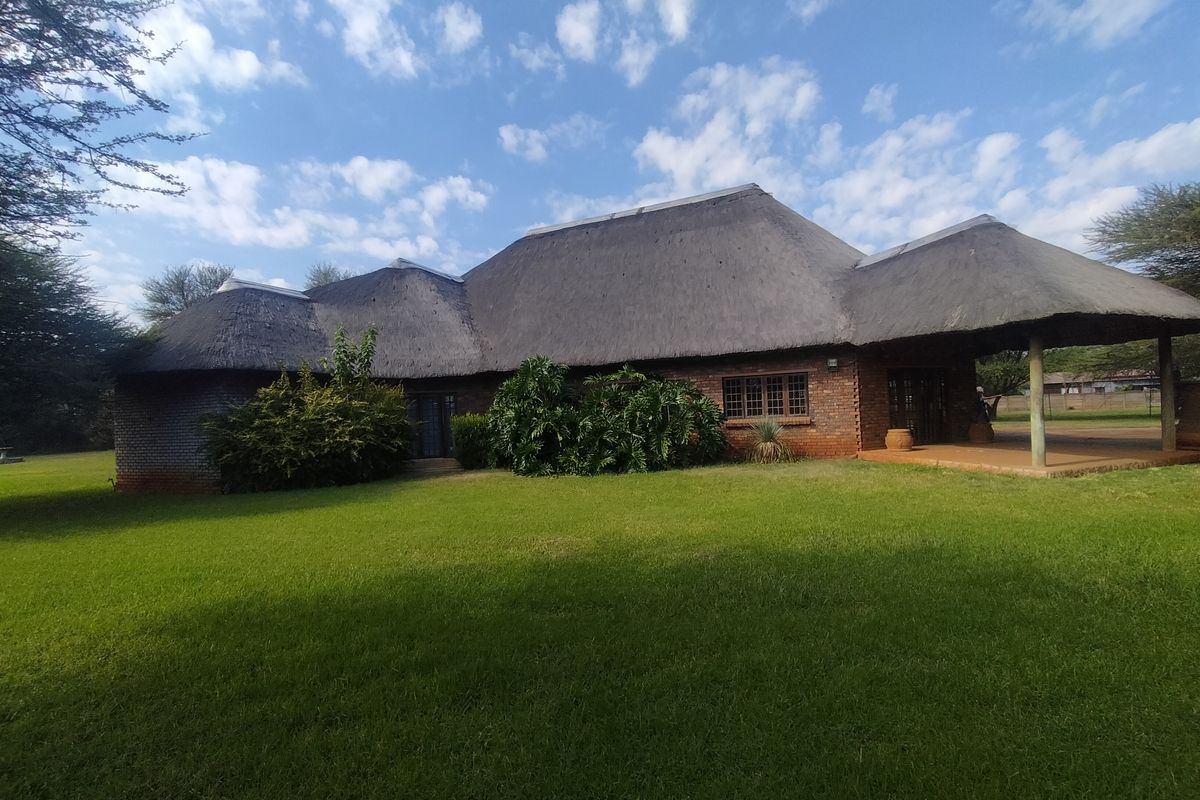 Rear view of a facebrick small holding residence featuring a traditional thatch roof and a spacious covered patio overlooking a large garden.