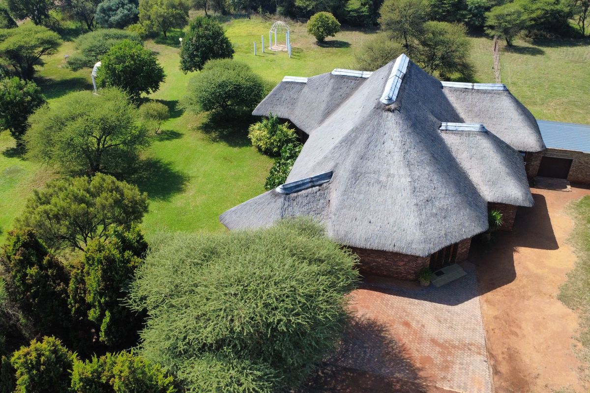 Aerial view of a property featuring a large thatch-roofed dwelling set within a spacious, well-maintained garden with mature trees and paved driveway.