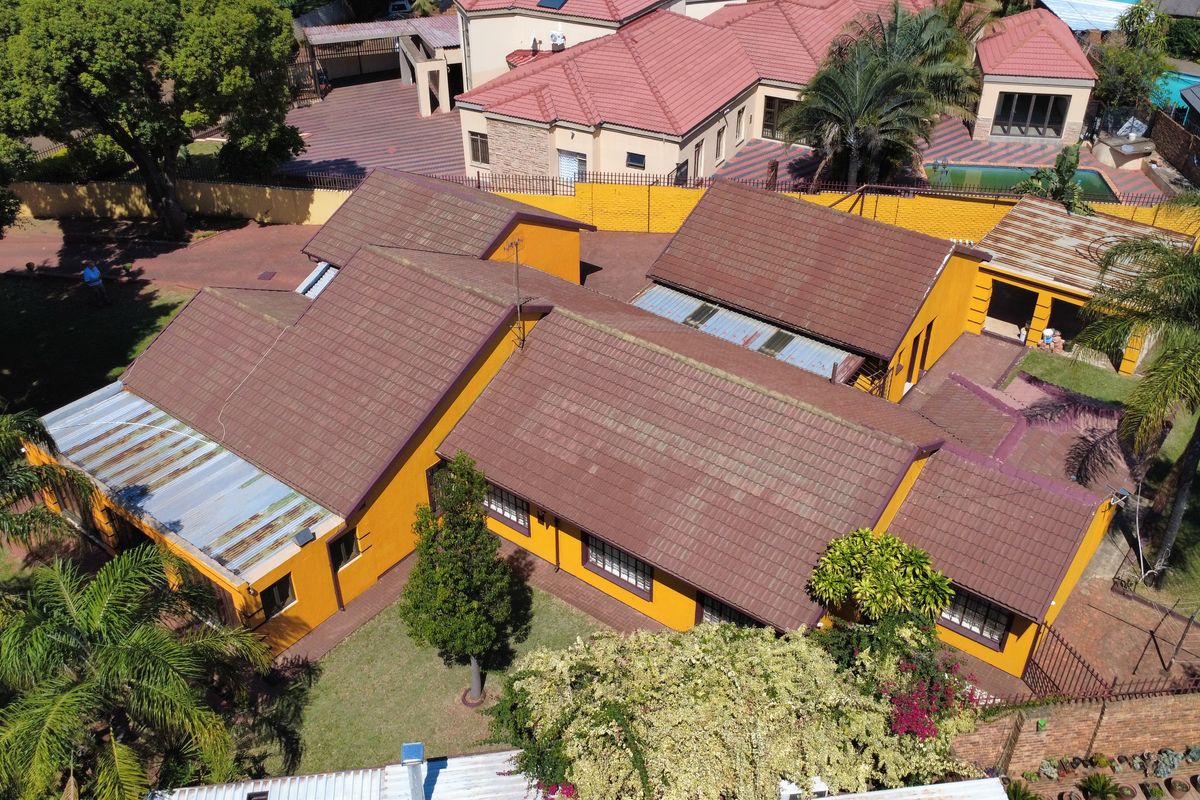 Aerial view of the property showcasing the main house with tiled and metal roofing, surrounding paved areas, and established garden landscaping.