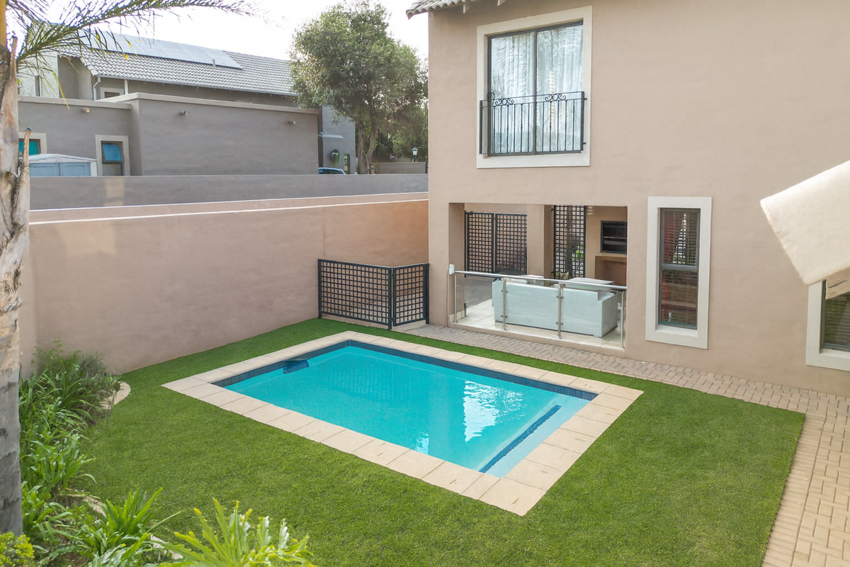 Rear exterior view featuring a swimming pool, artificial lawn, paved patio area, and a covered outdoor entertainment space with a built-in braai.