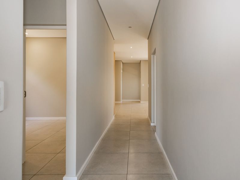 A clean, modern hallway featuring neutral wall tones, recessed downlighting, and large format floor tiles leading to adjacent rooms.