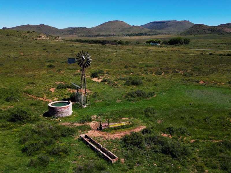 Aerial view of a rural farm landscape featuring a traditional windmill, a circular brick water reservoir, and livestock watering troughs.