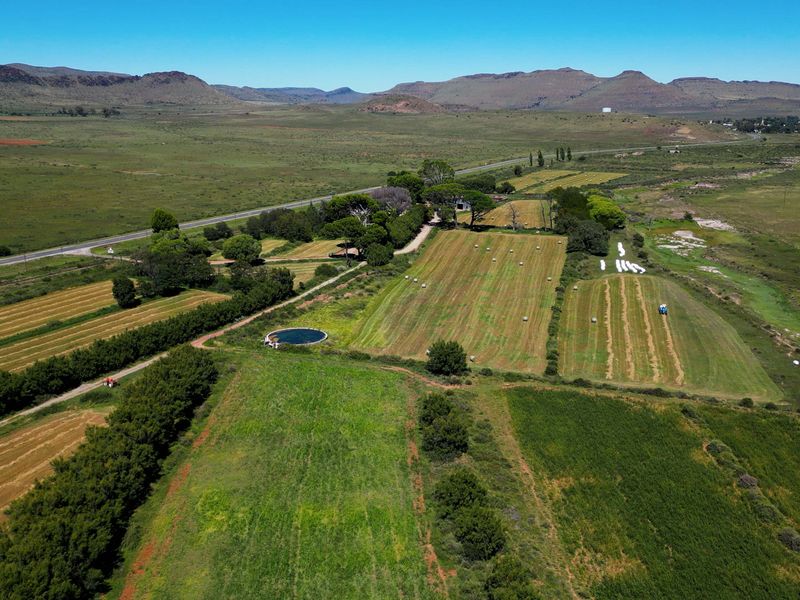 Aerial view of a vast farm property featuring expansive cultivated fields, mature trees, and a circular water reservoir set against a scenic mountain landscape.