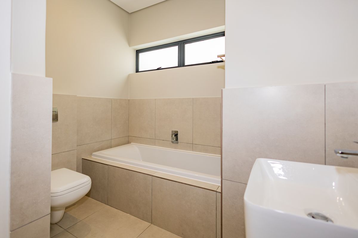 Modern bathroom featuring a built-in bathtub, wall-mounted toilet, and basin with neutral tiled finishes and a horizontal window for natural light.