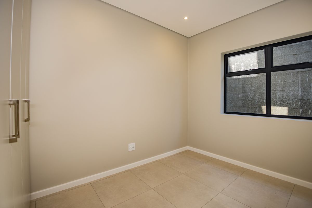 A clean, modern, and empty bedroom featuring neutral tiled flooring, built-in cupboards, and a dark aluminium window frame.