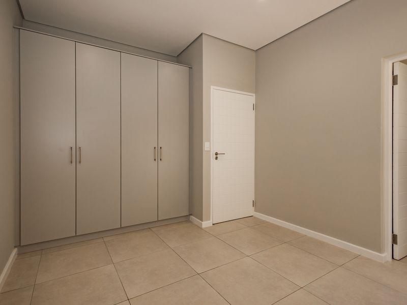 A clean, modern bedroom featuring neutral wall tones, large-format floor tiles, and extensive built-in cupboard storage.
