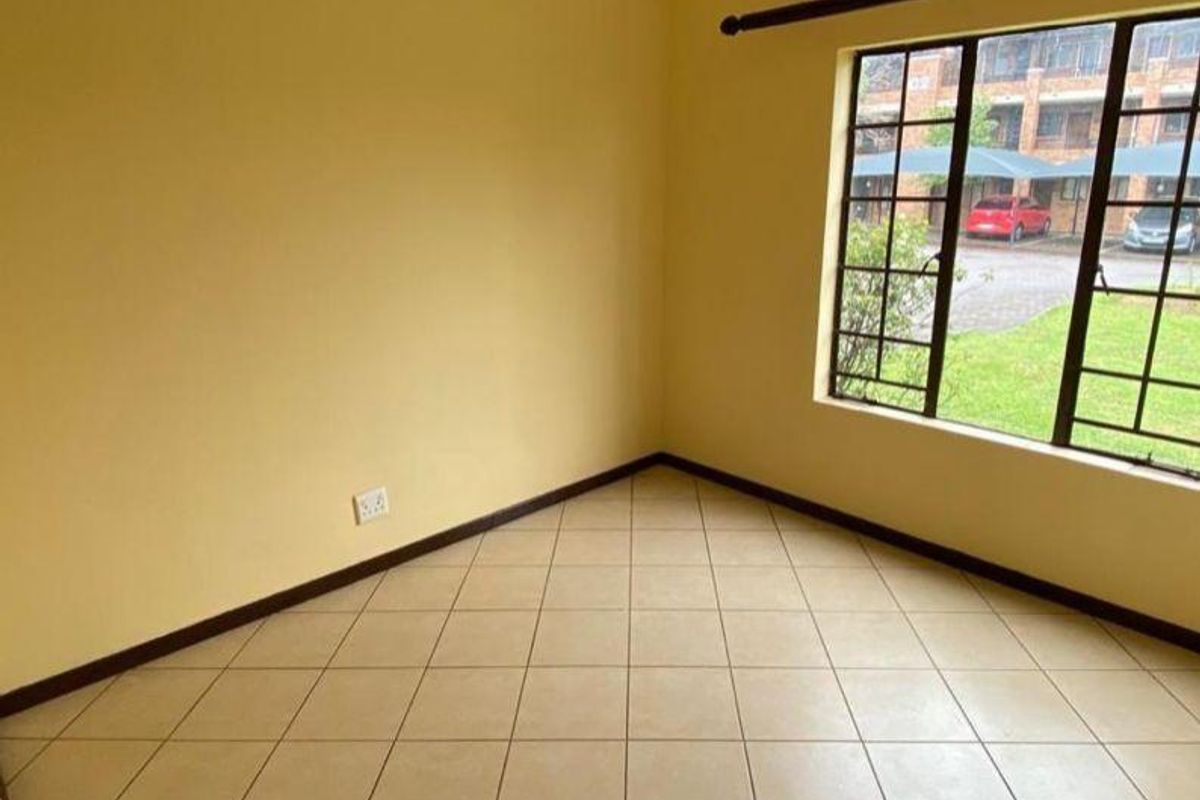 An empty bedroom featuring neutral yellow walls, tiled flooring, and a large metal-framed window overlooking the complex parking area.