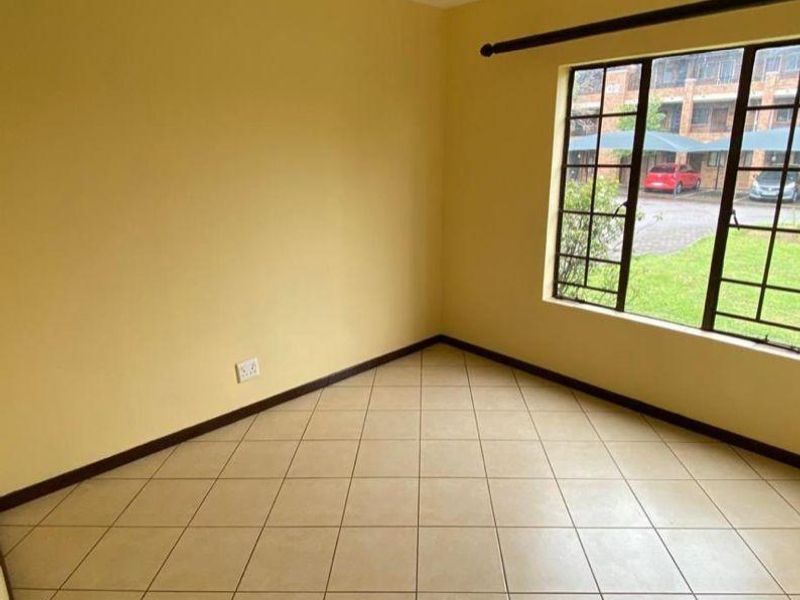 An empty bedroom featuring neutral yellow walls, tiled flooring, and a large metal-framed window overlooking the complex parking area.