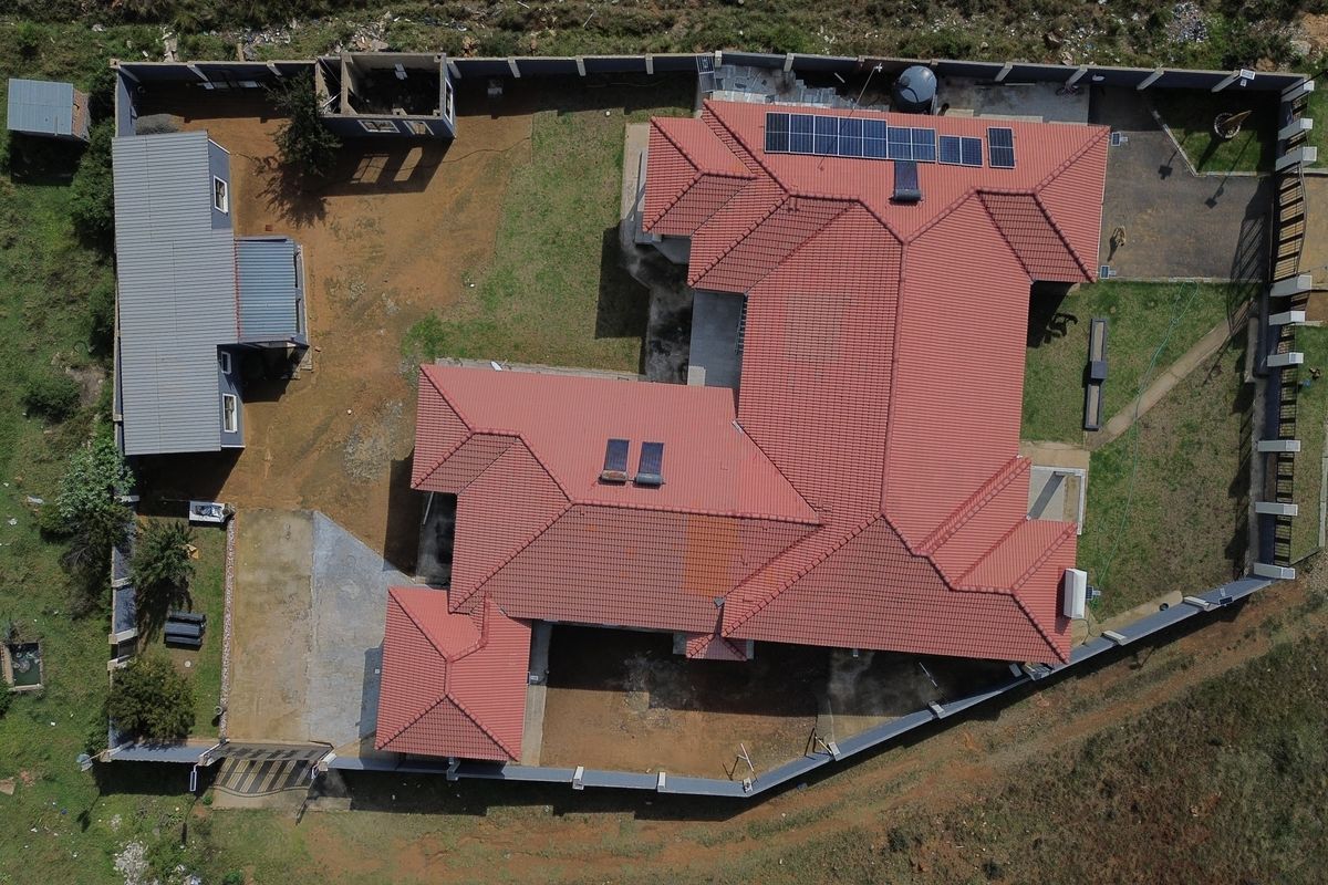 Aerial view of a residential property featuring a large tiled roof with solar installations, a perimeter wall, and an unfinished outbuilding structure.