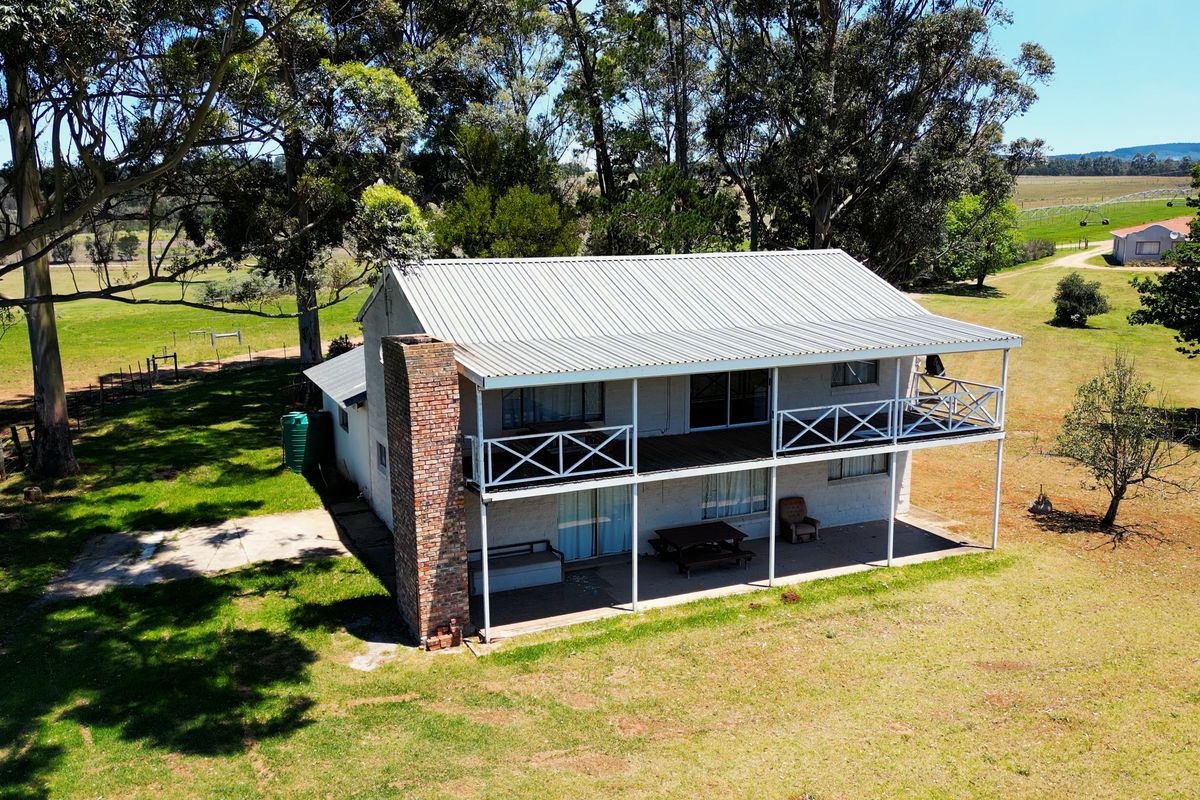 Aerial view of a two-story farmhouse featuring a corrugated metal roof, brick chimney, and a spacious wrap-around veranda set on a large rural property.