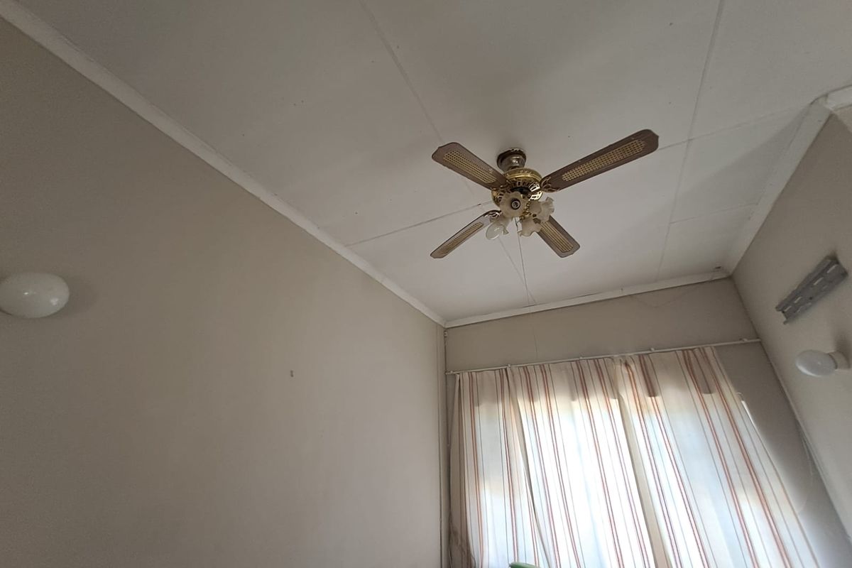 A bedroom interior featuring a ceiling fan and wall-mounted lighting, showing signs of wear on the ceiling panels.