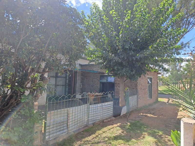 Front exterior view of a single-story exposed brick house featuring metal security fencing and mature trees in the front yard.