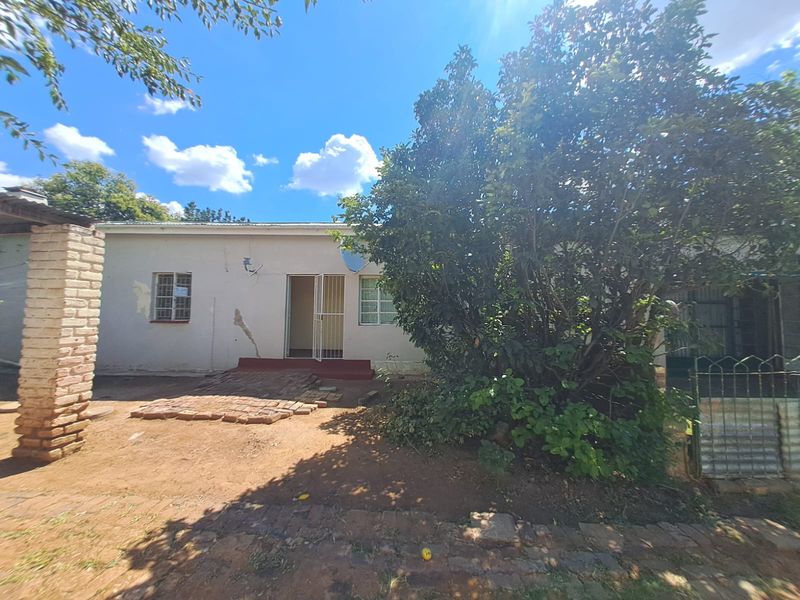 Rear view of the property showing a single-story structure with visible exterior wall damage and a large tree in the garden area.