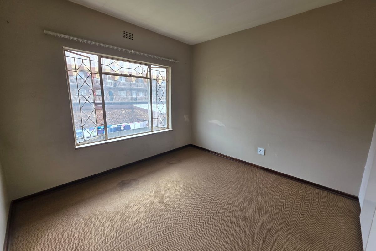 An empty bedroom featuring neutral walls, carpeted flooring, and a large window with security bars, offering a blank canvas for renovation.