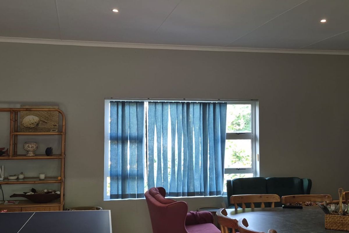 A spacious living area featuring neutral wall tones, recessed downlighting, and large windows fitted with vertical blinds.