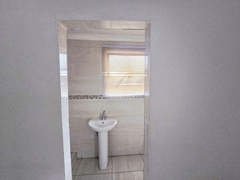 A compact bathroom featuring a pedestal basin, neutral wall and floor tiling, and a window with a fitted venetian blind.