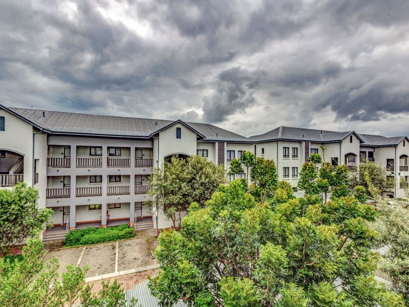 Exterior view of a multi-story apartment complex featuring paved parking areas, landscaped greenery, and covered balcony walkways.