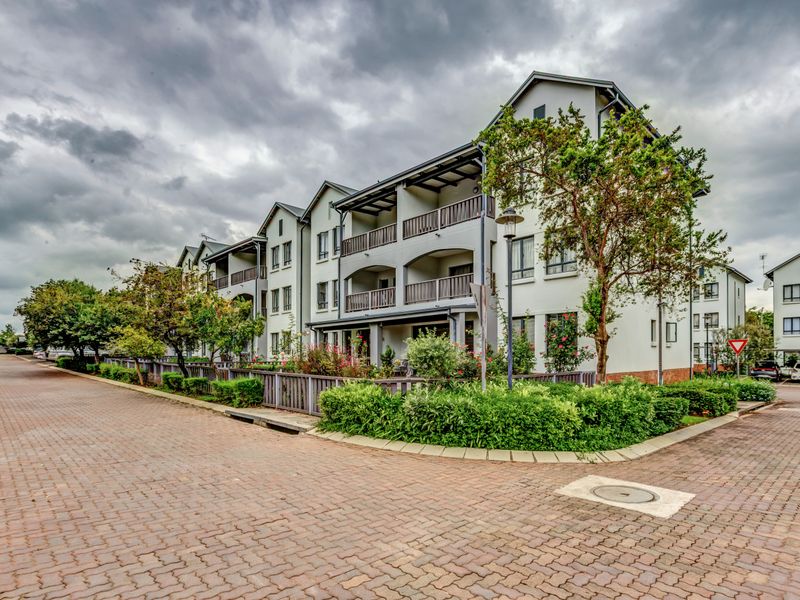 Exterior view of a multi-story apartment complex featuring paved walkways, landscaped garden beds, and private balconies with wooden railings.