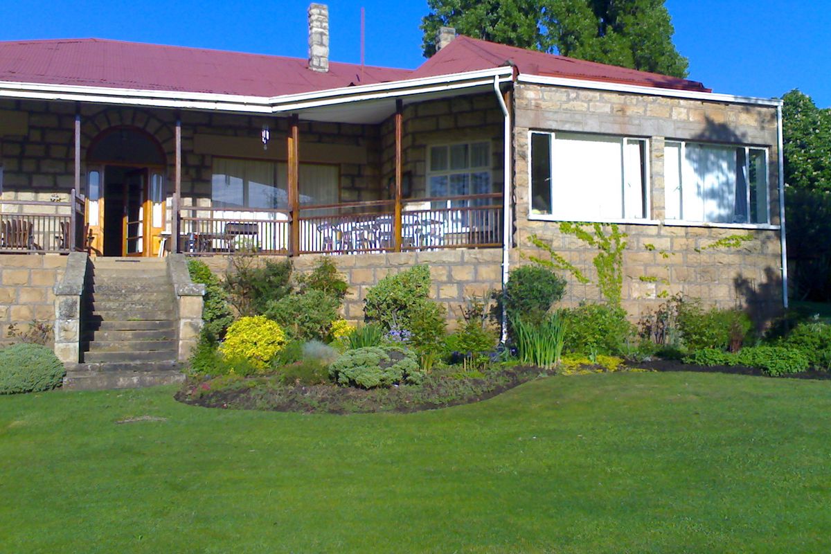 A charming stone-clad farmhouse featuring a covered veranda, red metal roof, and well-maintained garden with stone steps leading to the entrance.