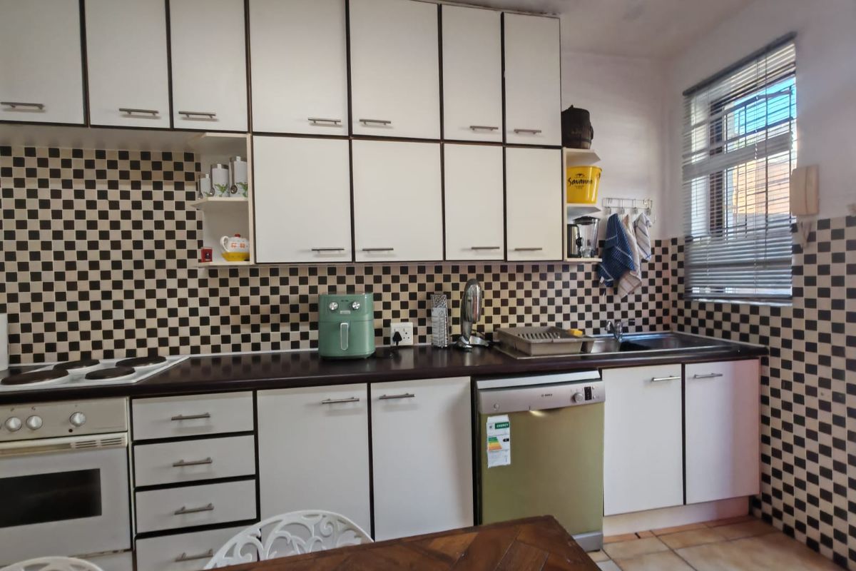 A functional kitchen featuring white cabinetry, dark laminate countertops, a checkered tile backsplash, and integrated appliances, showing signs of age and wear.