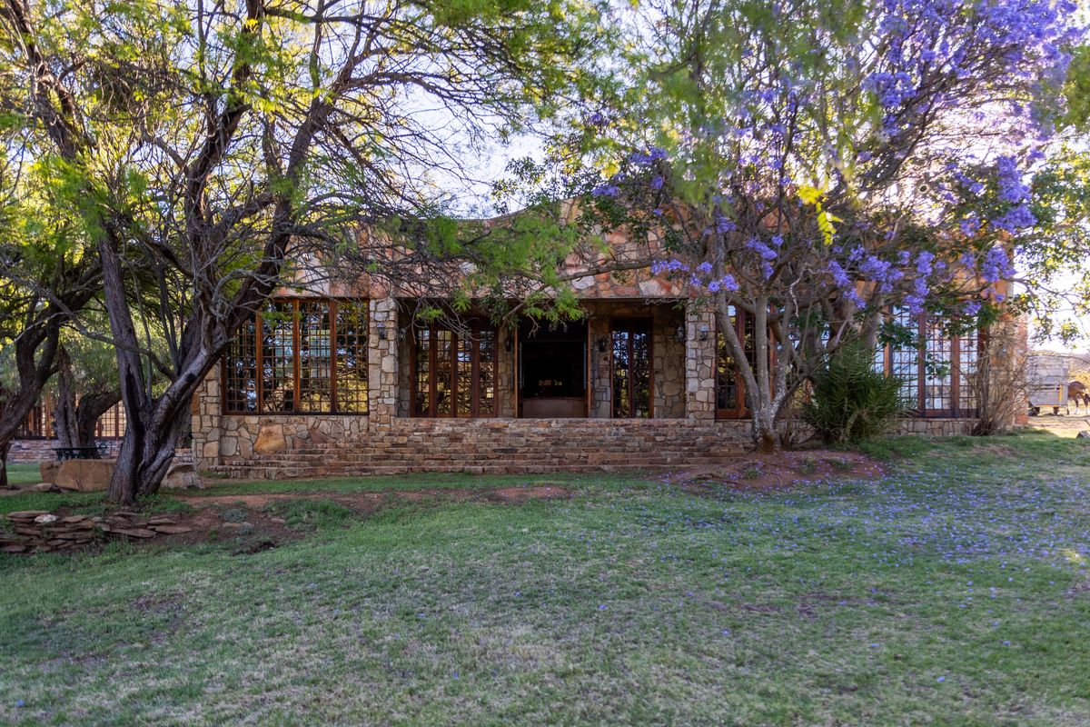 A charming stone-clad farmhouse exterior featuring large timber-framed windows and a covered patio area, set within a mature garden with established trees.