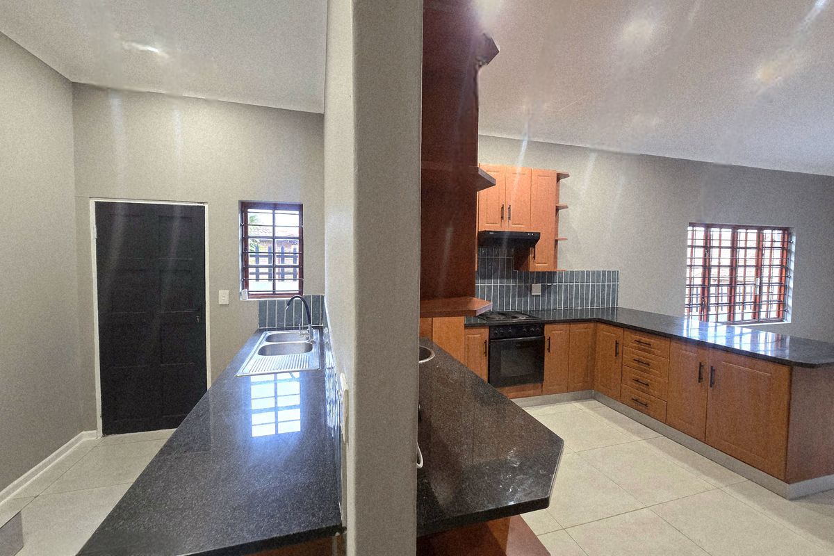 A spacious, empty kitchen featuring wooden cabinetry, dark granite countertops, built-in oven and hob, and tiled flooring with a separate scullery area.