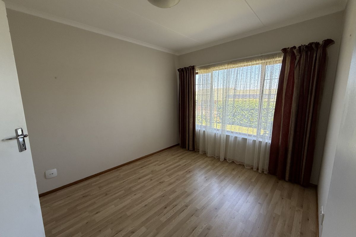 A bright, empty bedroom featuring light-toned laminate flooring, neutral walls, and a large window with curtain fixtures.
