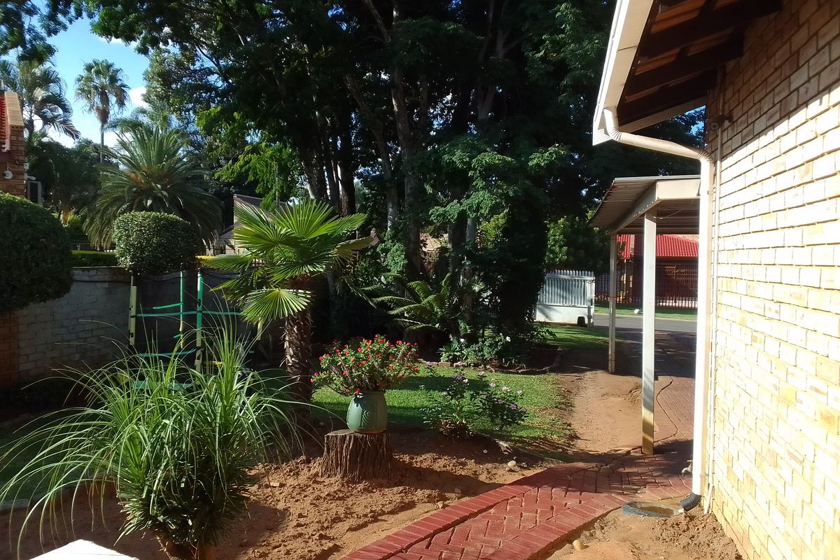 A view of the rear garden area featuring a brick pathway, established trees, and a covered patio section attached to the main house structure.