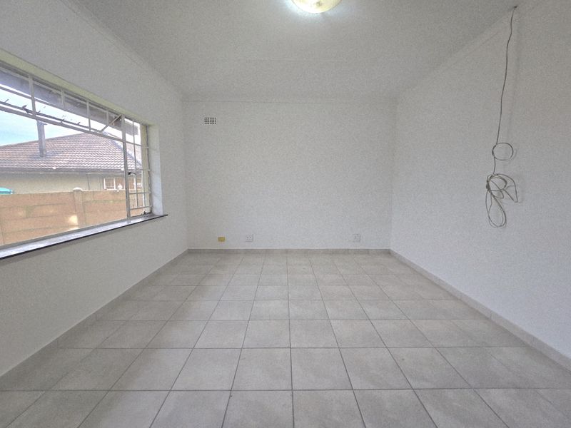 A vacant, neutrally finished room featuring tiled flooring, a large window with security bars, and exposed electrical wiring.