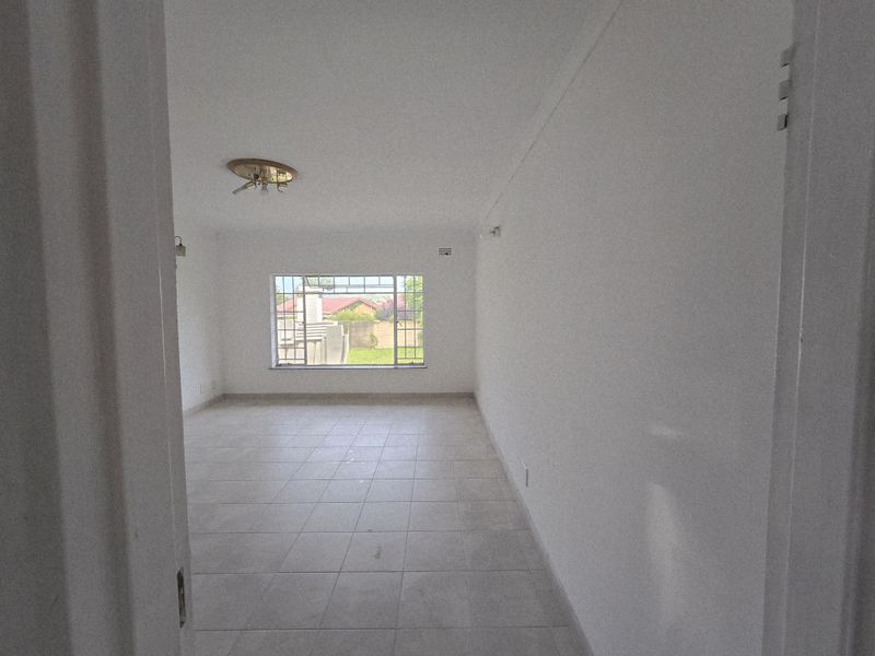 A bright, empty living room featuring neutral tiled flooring, a large window with security bars, and a simple ceiling light fixture.