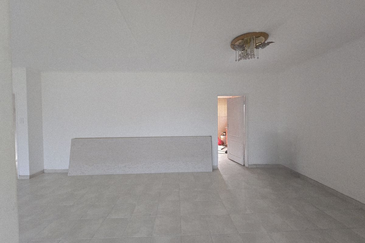 A spacious, empty living area featuring neutral floor tiling, a central ceiling light fixture, and a doorway leading to an adjacent room.