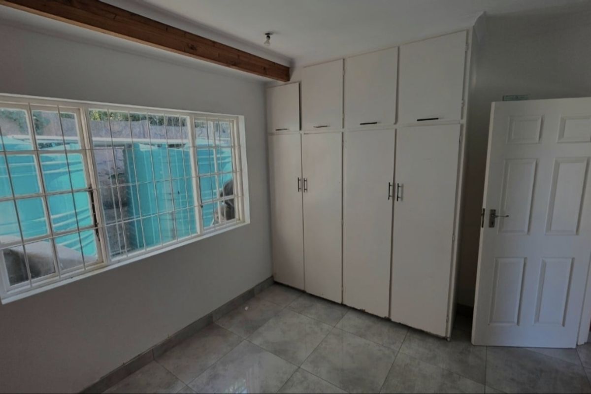 A clean, empty bedroom featuring tiled flooring, built-in cupboards for storage, and a window fitted with security bars.
