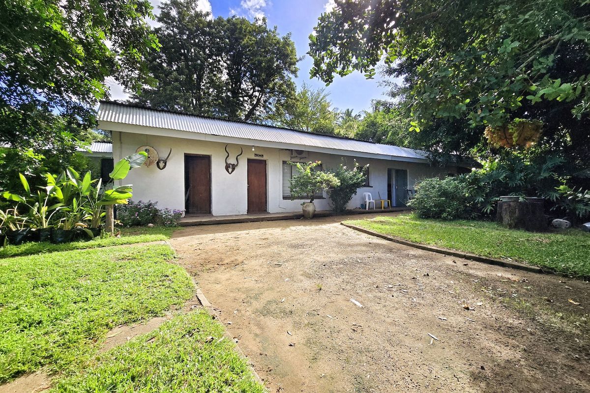 A single-story exterior view of a small holding dwelling featuring a corrugated metal roof, multiple entrance doors, and a surrounding garden area.