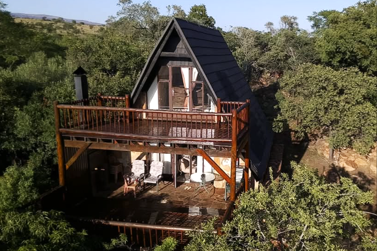 An elevated view of an A-frame townhouse featuring a spacious timber deck and balcony, nestled within a natural, wooded environment.