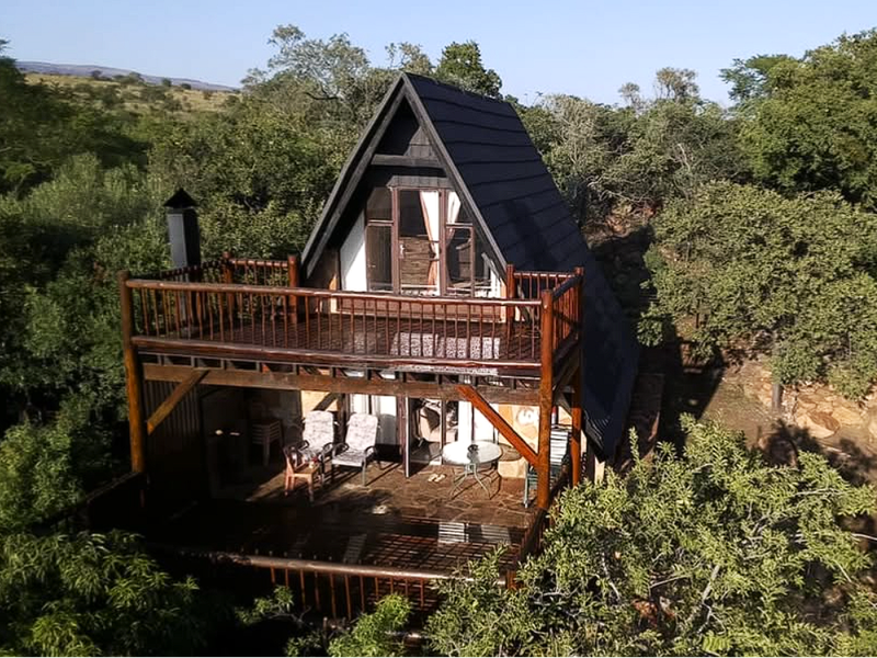 An elevated view of an A-frame townhouse featuring a spacious timber deck and balcony, nestled within a natural, wooded environment.