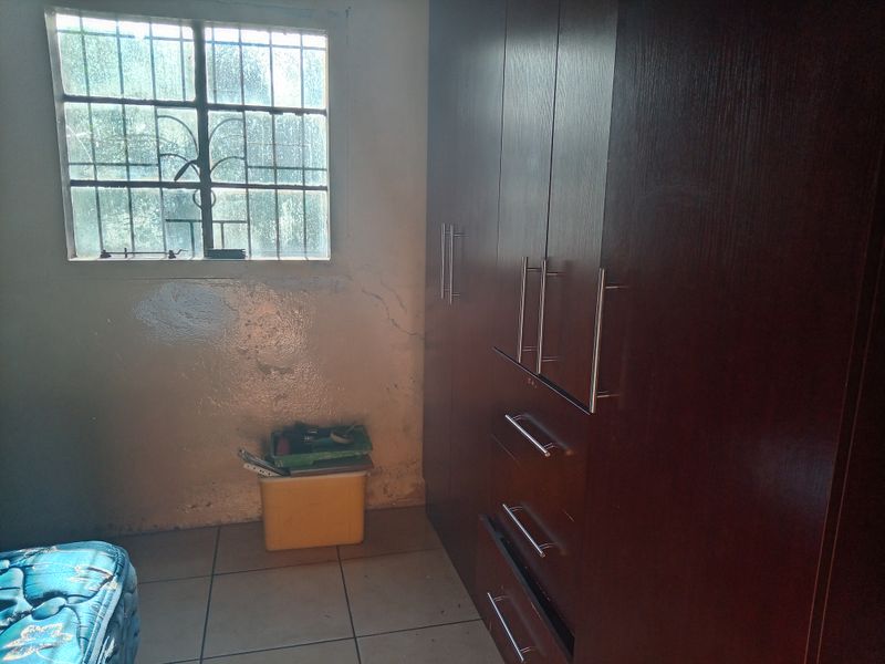 A bedroom featuring built-in cupboards, tiled flooring, and a window with security bars. The walls show signs of wear and require maintenance.