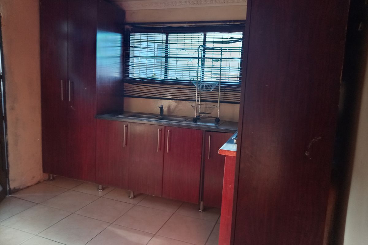 A compact kitchen featuring dark wood-finish cabinetry, laminate countertops, a stainless steel sink, and tiled flooring, requiring cosmetic updates to improve its overall appeal.