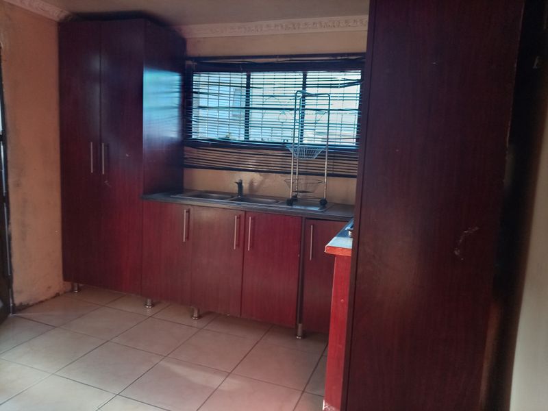 A compact kitchen featuring dark wood-finish cabinetry, laminate countertops, a stainless steel sink, and tiled flooring, requiring cosmetic updates to improve its overall appeal.