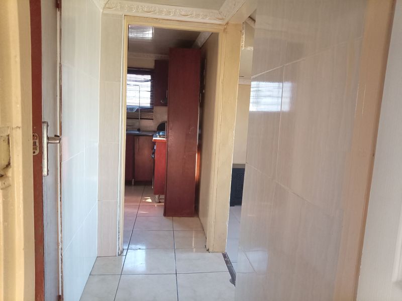 A narrow hallway with tiled walls and flooring, leading into a kitchen area, featuring crown molding and dated finishes.